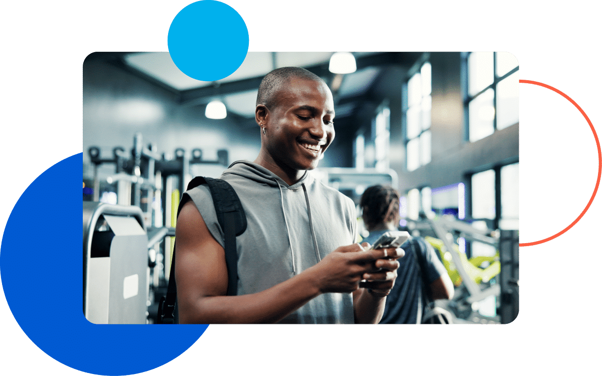What is Pay Day Graphic Man smiling and using a smartphone at the gym with modern decor in the background.