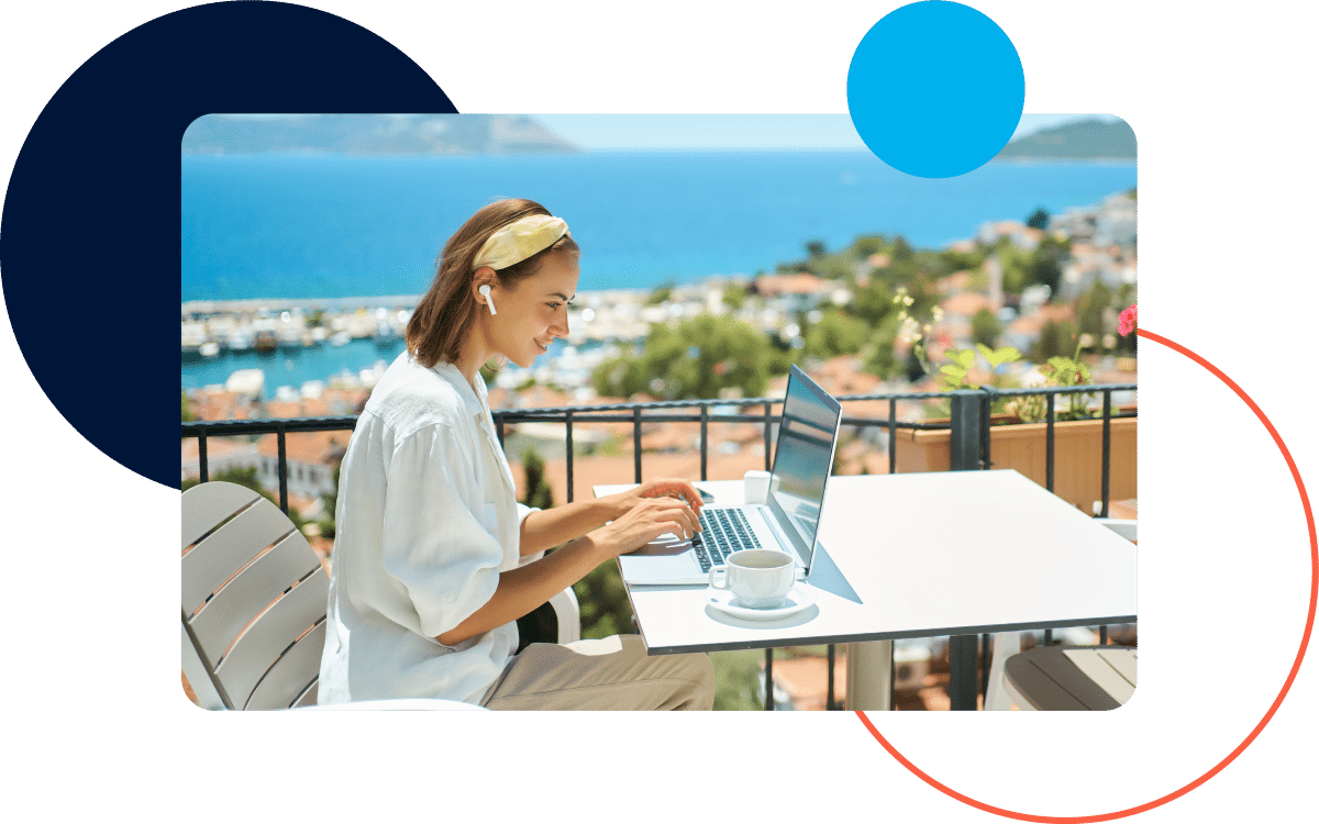 WhoCanUseGraphics Woman working on a laptop outdoors with a cup of coffee, overlooking a coastal town.